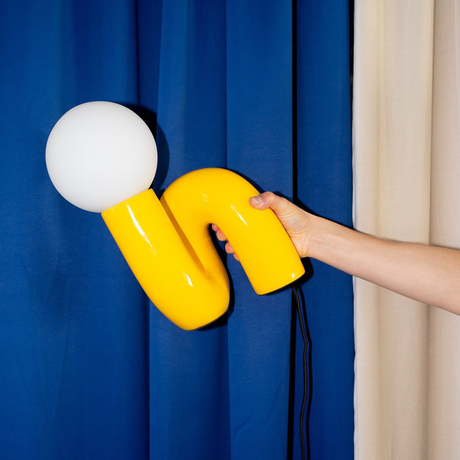 Gelbe Snake Lampe in der Hand gehalten vor königsblauem Vorhang – geschwungene Retro-Form mit runder Glaskugel und schwarzem Kabel