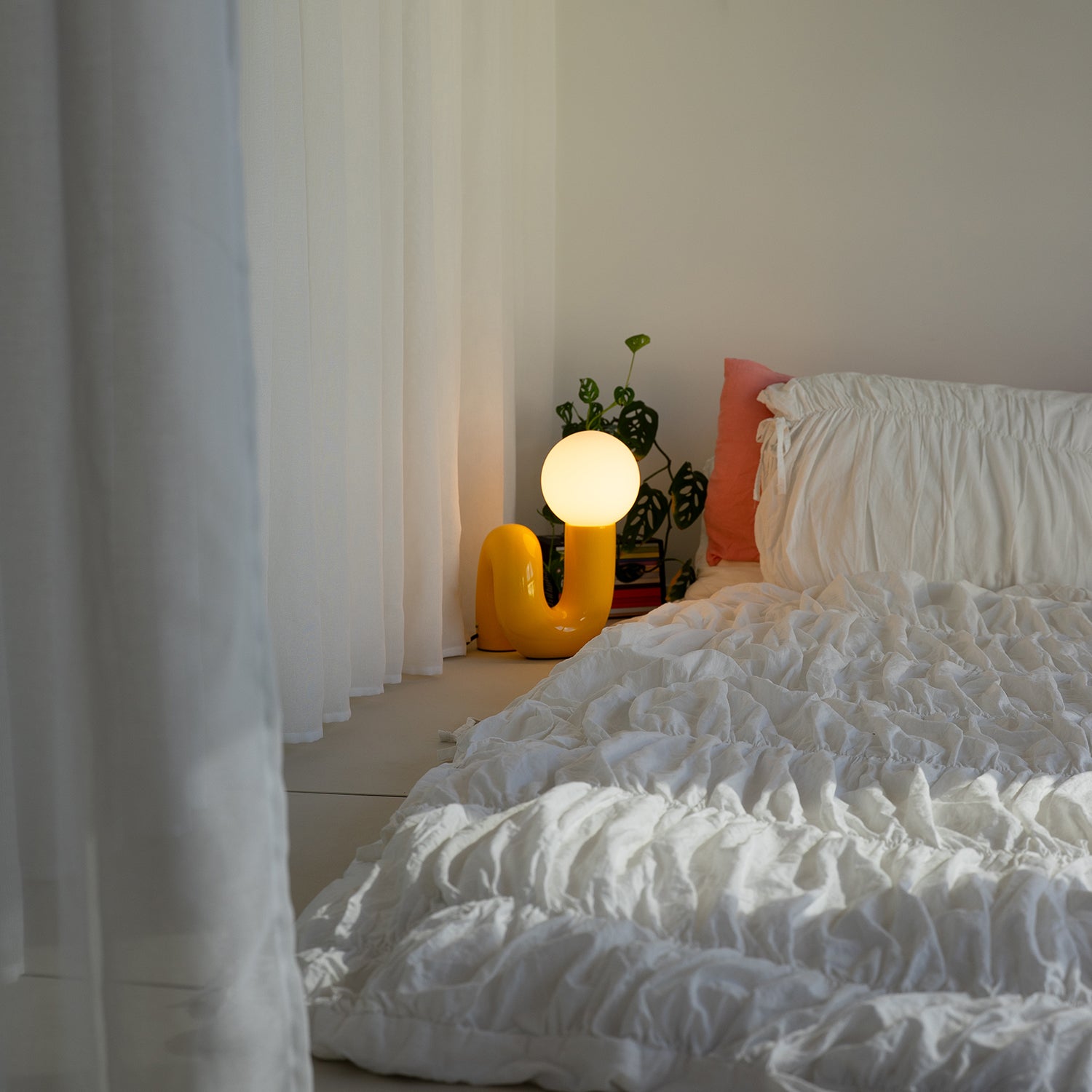 Gelbe Snake Lampe als Nachttischlampe neben Bett mit weißer Leinenbettwäsche und Monstera-Pflanze im minimalistischen Schlafzimmer