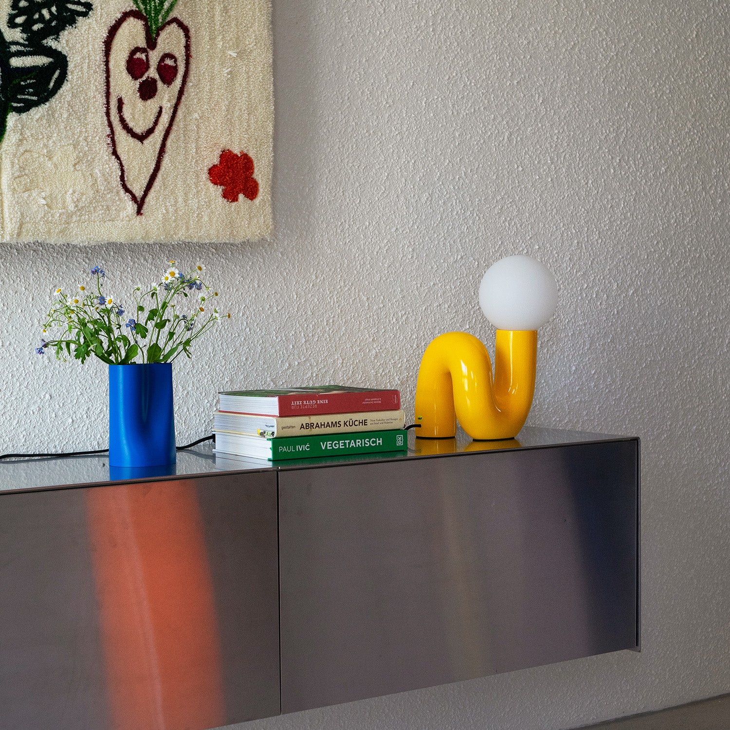 Gelbe Snake Tischlampe auf dunklem Metall-Sideboard neben Büchern und blauer Vase – 60er Retro Design im Wohnzimmer-Setting