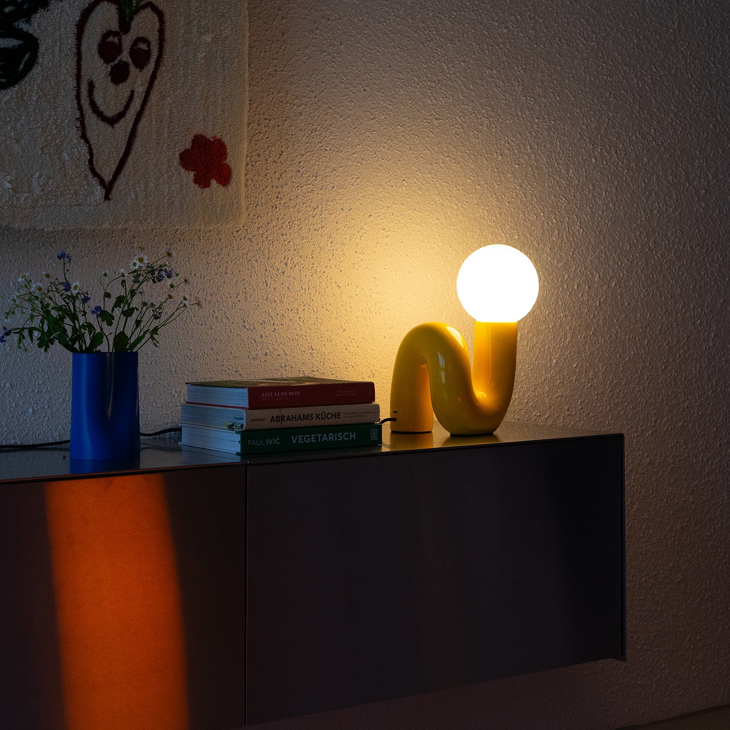 Gelbe Snake Lampe leuchtend auf Sideboard mit warmem Abendlicht – stimmungsvolle Retro-Szene mit Wandbehang und Blumen