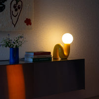 Gelbe Snake Lampe leuchtend auf Sideboard mit warmem Abendlicht – stimmungsvolle Retro-Szene mit Wandbehang und Blumen