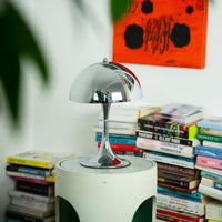 Eine glänzende Chromlampe im Vintage Retro Stil steht auf einem Tisch, umgeben von einer Sammlung Bücher. Diese elegante Tischlampe, inspiriert von der Space Age Ära, ist ideal für das Arbeitszimmer oder als stilvolle Raumbeleuchtung.