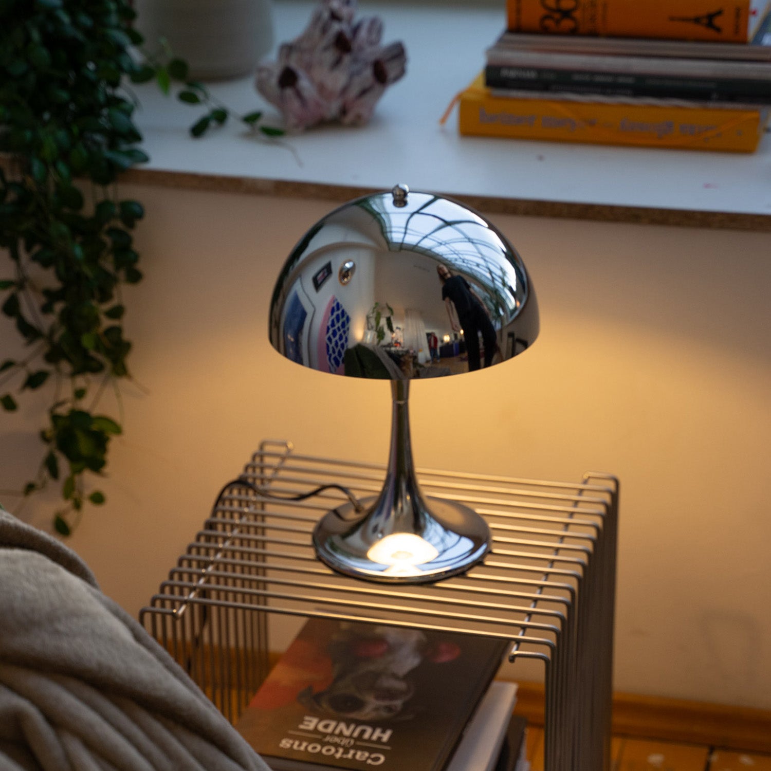 Chrome Bell von GLOW UP STUDIO – eine glänzend verchromte Retro-Tischlampe im ikonischen Mid Century Look. Die halbrunde Glockenform reflektiert ihre Umgebung wie ein Spiegel und steht auf einem eleganten, trompetenförmigen Fuß. Die Lampe steht auf einem modernen Gittertisch unter einem Fensterbrett mit Pflanzen und Kunstbüchern. Ihr warmes Licht sorgt für gemütliche Stimmung im Raum – ein stilvoller Eyecatcher mit Space Age Vibes.