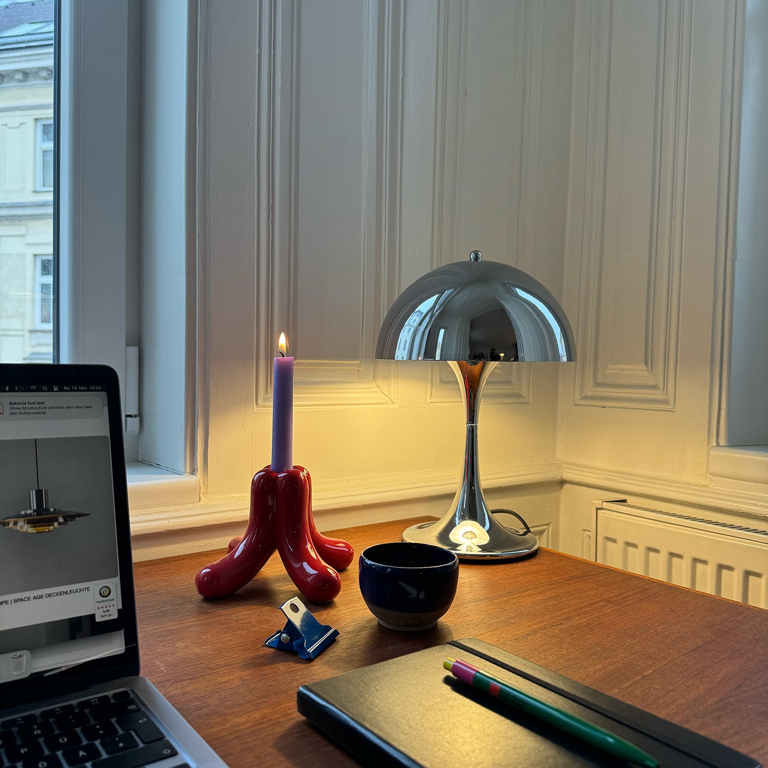 Zeitlose Chrome Lampe im Retro Design von GLOW UP STUDIO auf Schreibtisch mit glänzender Retro-Tischlampe, brennender lilafarbener Kerze in rotem Kerzenhalter, Laptop, Notizbuch mit bunten Stiften und kleiner Tasse. Warmes, gemütliches Licht vor einer weißen Wand mit Fenster.