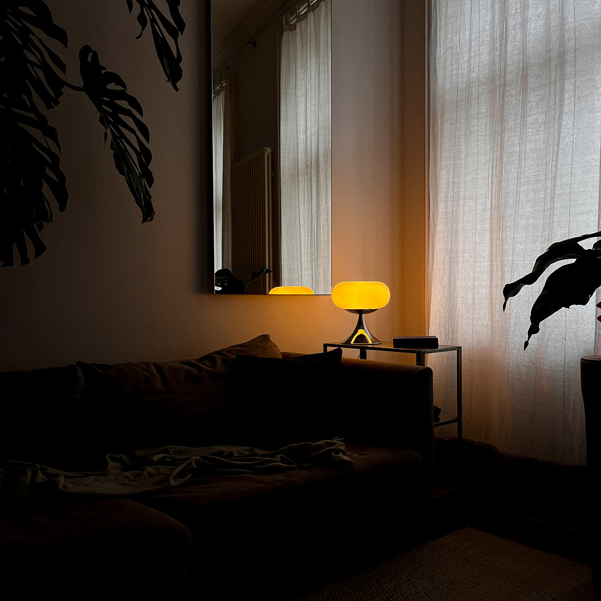 Die Mid-Century Lampe verströmt ein weiches, warmes Licht. Sie steht auf einem modernen Beistelltisch neben einem dunkelgrauen Sofa, das mit einer Decke bedeckt ist. Große Monstera-Blätter werfen Schatten auf die Wand, die Vorhänge sind zugezogen.