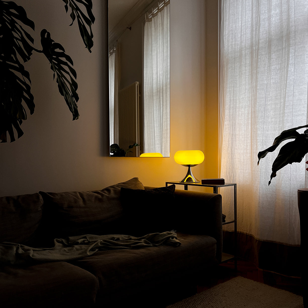 Die eingeschaltete Space Age Lampe taucht den Raum in warmes, weiches Licht. Sie steht auf einem Metall-Beistelltisch, spiegelt sich im großen Wandspiegel. Eine große Monstera-Pflanze wirft Schatten auf die Wand, die weißen Vorhänge sind leicht zugezogen. Das dunkle Sofa mit einer locker drapierten Decke sorgt für eine gemütliche, abendliche Atmosphäre.