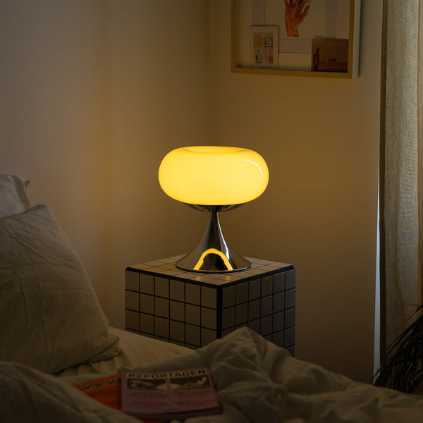 Die Space Age Lampe strahlt warmes Licht aus, platziert auf einem karierten Beistelltisch neben einem Bett. Die Bettdecke ist leicht zerknittert, Zeitschriften liegen darauf. Die Atmosphäre ist ruhig und entspannend, perfekt für den Abend.