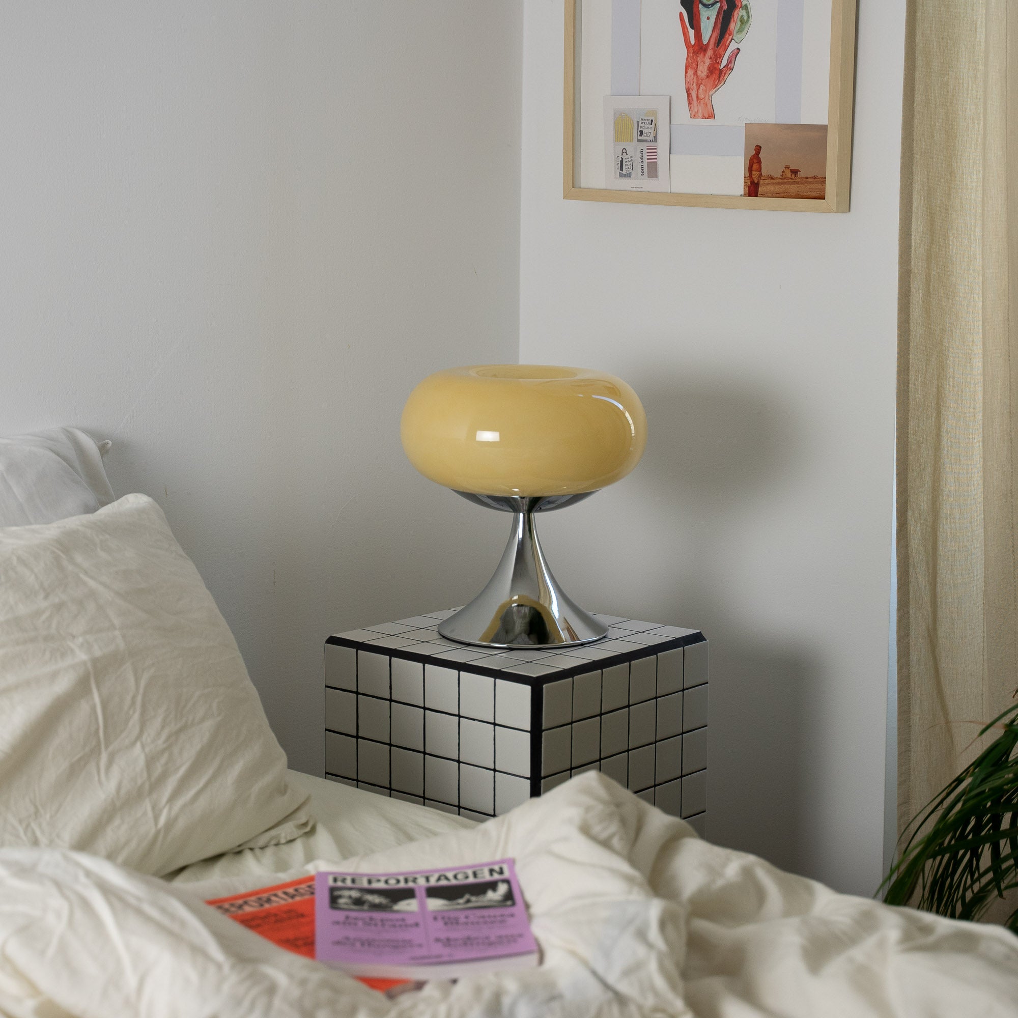 Cremefarbene Space Age Tischlampe auf einem karierten Nachttisch neben einem Bett mit beiger Bettwäsche. Sanfte, warme Farbtöne dominieren den Raum. An der Wand hängen Collagen und Kunstprints, die den gemütlichen Charakter unterstreichen.