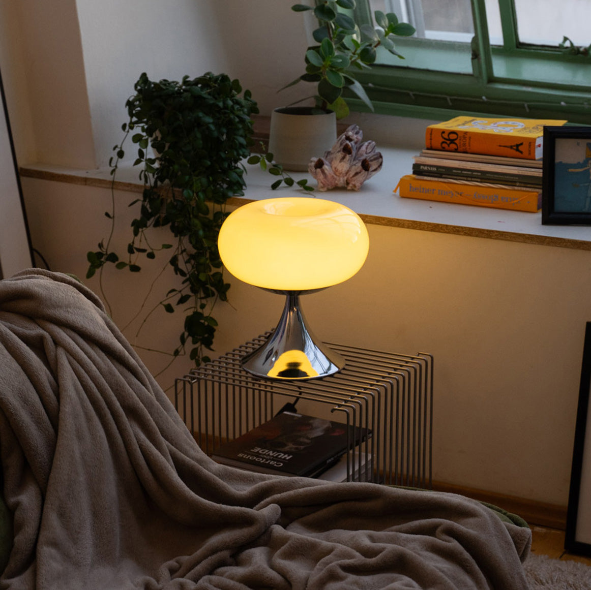 Gelbe Donut Lampe von GLOW UP STUDIO auf einem modernen Draht-Beistelltisch vor einer Fensterbank mit Pflanzen, Büchern und Dekoobjekten. Die Lampe mit ihrem leuchtenden, runden Glasschirm und dem verchromten Fuß sorgt für ein warmes, gemütliches Licht. Im Vordergrund liegt eine weiche beigefarbene Decke auf einem Sofa – das perfekte Setting für ein hyggeliges Wohnzimmer mit Retro-Charme und Mid Century Vibes.