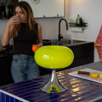 Grüne Donut Lampe von GLOW UP STUDIO auf einer dunkelblau gefliesten Kücheninsel mit glänzendem Chromfuß – ein echtes Space Age Designstück im Retro-Stil. Im Hintergrund steht eine junge Frau mit Kaffee und Orange in der Hand in einer modernen, minimalistischen Küche. Die Szene wirkt lebendig und modern – perfekt für alle, die auffällige Mid Century Eyecatcher in ihr Zuhause bringen wollen.