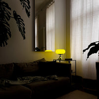 Die Retro-Lampe taucht den Raum in warmes Licht, reflektiert sich im Spiegel und sorgt für eine stimmungsvolle Beleuchtung. Ein großes Sofakissen, eine Monstera-Pflanze und weiche Vorhänge unterstreichen die gemütliche Abendstimmung.