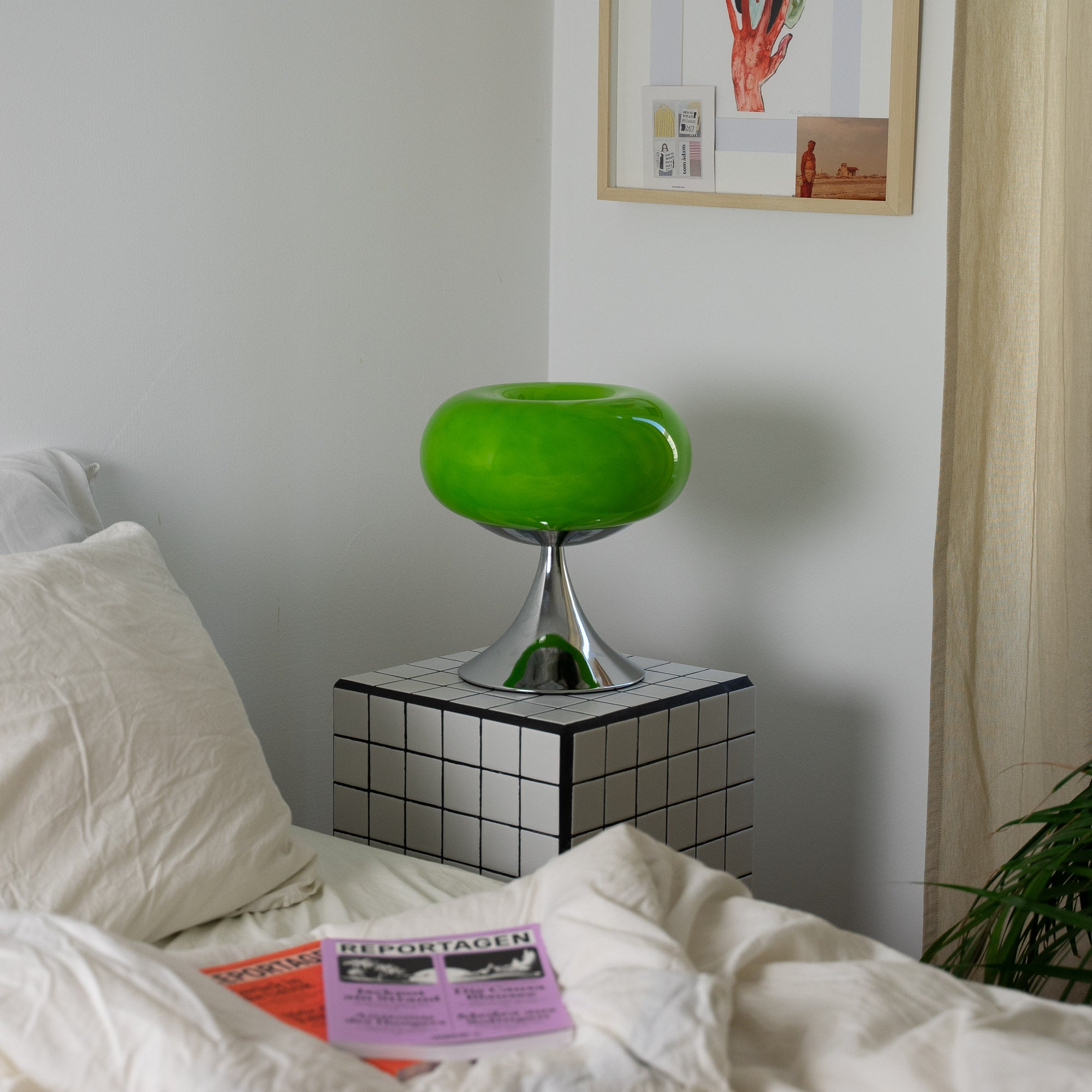 Die grüne Mid-Century-Lampe auf einem karierten Beistelltisch neben einem Bett mit beiger Bettwäsche. Eine Zeitschrift liegt auf der Decke, sanftes Tageslicht fällt durch helle Vorhänge und verleiht dem Raum eine ruhige Atmosphäre.