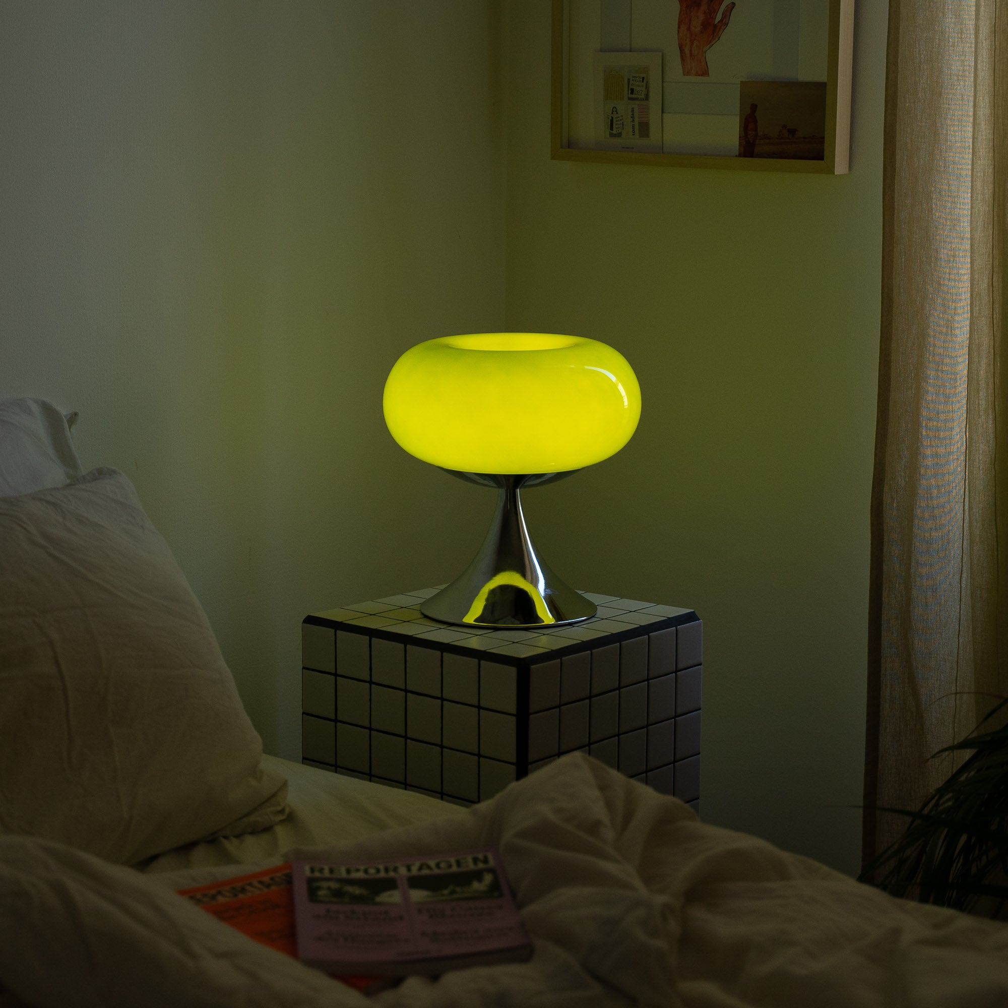 Die grüne Space Age Lampe verbreitet ein warmes, beruhigendes Licht. Die Kombination aus kariertem Beistelltisch, beiger Bettwäsche und sanften Vorhängen sorgt für eine gemütliche Schlafzimmereinrichtung.