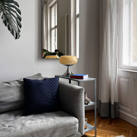 Die cremefarbene Designlampe mit glänzendem Chromfuß steht auf einem minimalistischen Metall-Beistelltisch neben einem grauen Sofa mit strukturiertem Stoff. Ein dunkelblaues Kissen setzt einen Kontrast. Helles Tageslicht fällt durch hohe Fenster mit weißen Vorhängen.