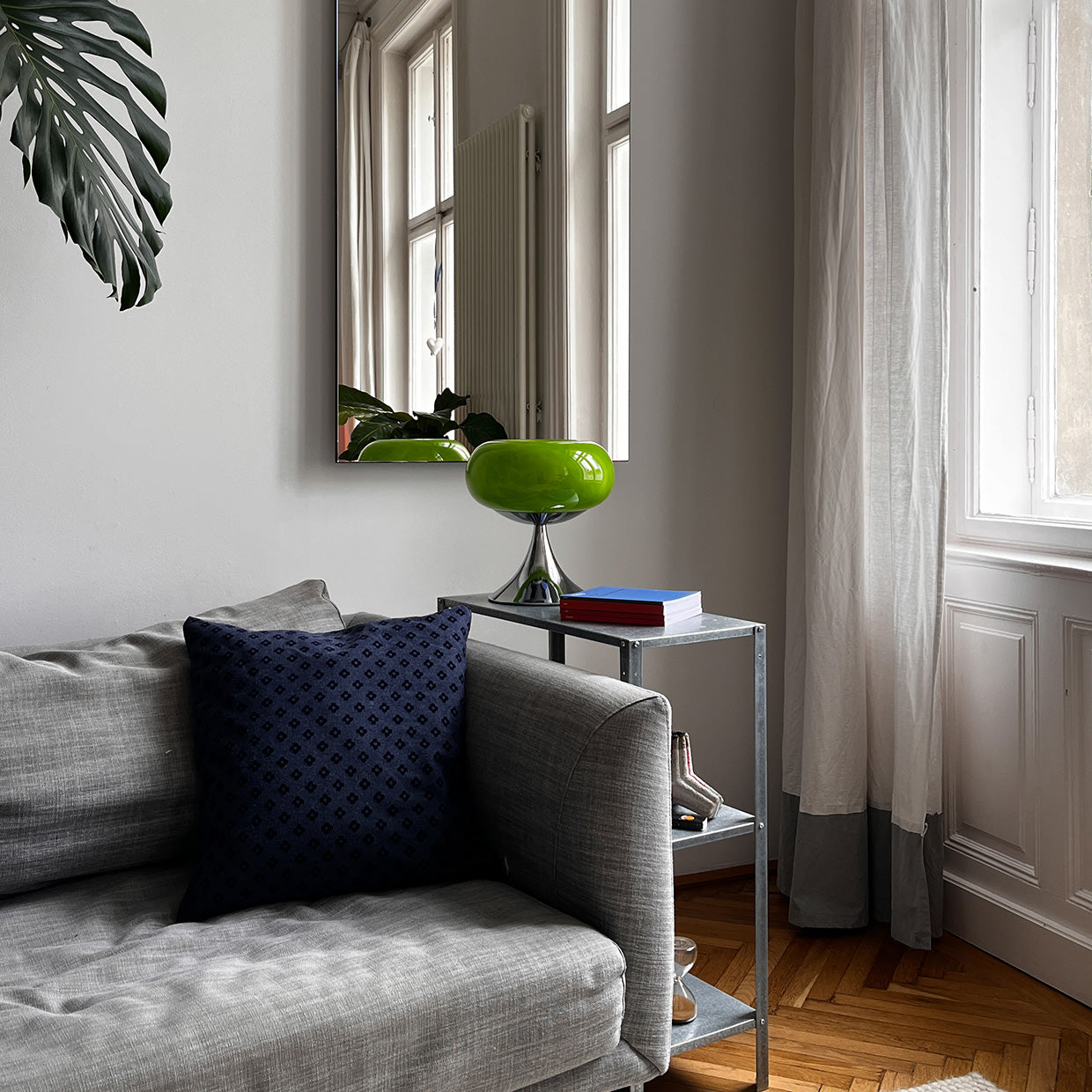 Die Tischlampe mit glänzendem Chromfuß steht auf einem modernen Metall-Beistelltisch neben einem grauen Sofa. Ein dunkelblaues Kissen setzt einen starken Kontrast, während Tageslicht durch die weißen Vorhänge fällt.