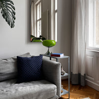 Die Tischlampe mit glänzendem Chromfuß steht auf einem modernen Metall-Beistelltisch neben einem grauen Sofa. Ein dunkelblaues Kissen setzt einen starken Kontrast, während Tageslicht durch die weißen Vorhänge fällt.