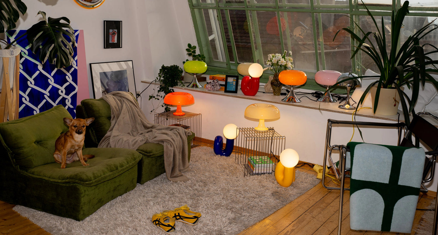Gemütliches Wohnzimmer mit grünem Retro-Sessel, auf dem ein brauner Hund sitzt. Große Fensterbank mit bunten Midcentury-Lampen, Pflanzen und Blumen. Teppich, Wolldecke, gelbe Hausschuhe und Stuhl mit grüner Decke runden die warme Atmosphäre ab.