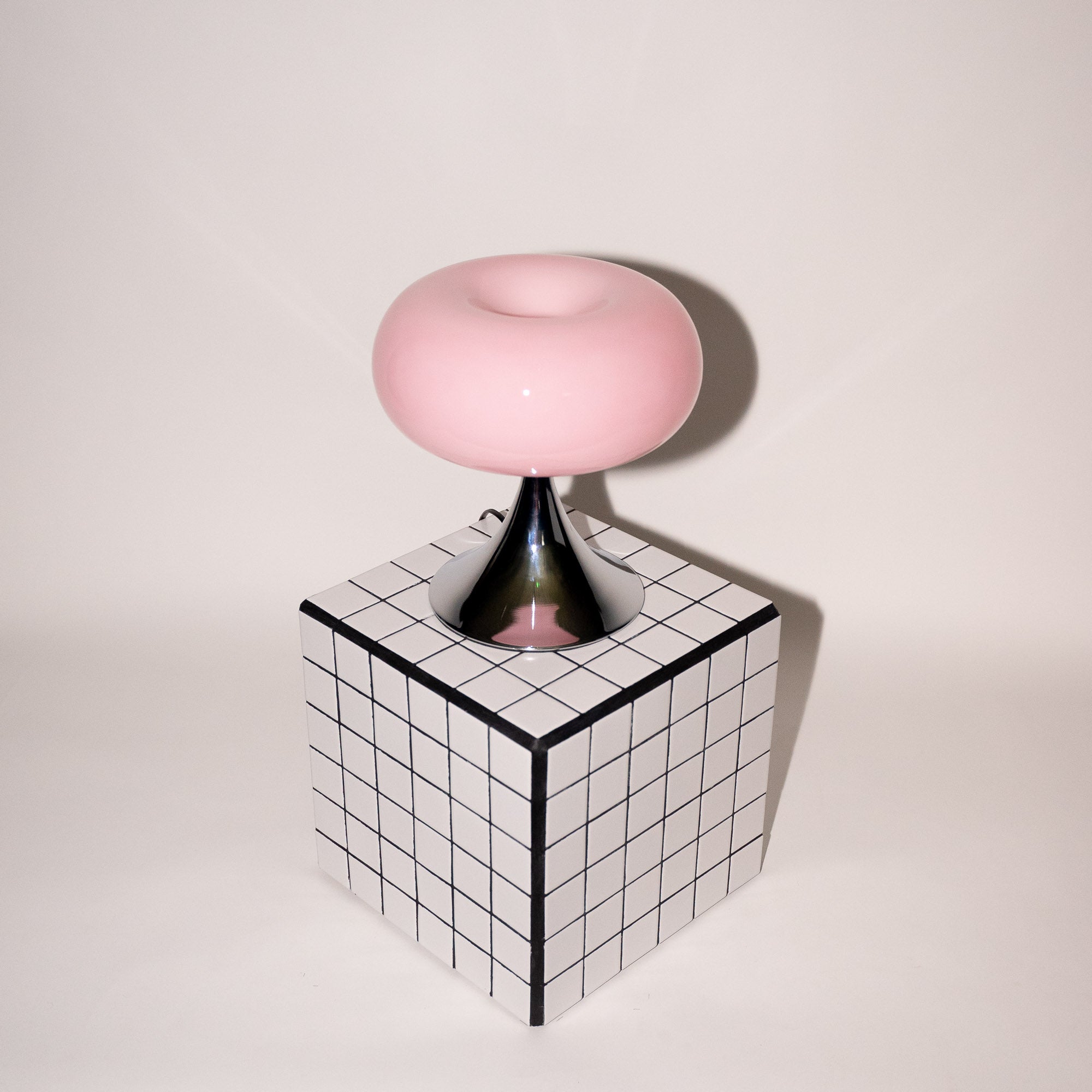 Sanft rosa Glasleuchte mit glänzendem Chromfuß, stilvoll platziert auf einem schwarz-weiß karierten Würfel. Das verspielte Design vereint 60er-Jahre-Ästhetik mit modernem Minimalismus.