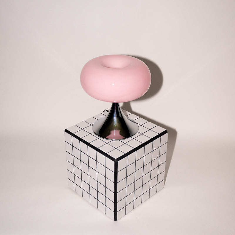 Sanft rosa Glasleuchte mit glänzendem Chromfuß, stilvoll platziert auf einem schwarz-weiß karierten Würfel. Das verspielte Design vereint 60er-Jahre-Ästhetik mit modernem Minimalismus.