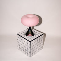 Sanft rosa Glasleuchte mit glänzendem Chromfuß, stilvoll platziert auf einem schwarz-weiß karierten Würfel. Das verspielte Design vereint 60er-Jahre-Ästhetik mit modernem Minimalismus.