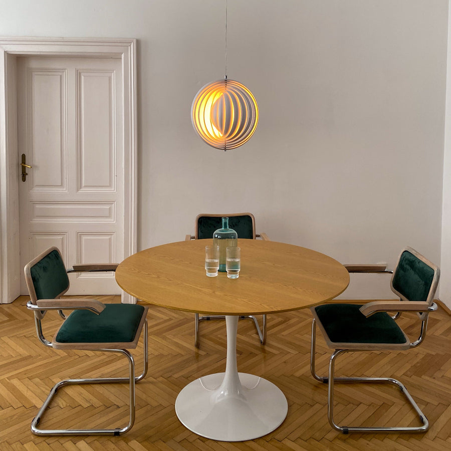 Pendelleuchte im Space Age Design über einem runden Holztisch mit weißem Tulpenfuß. Umgeben von vier Freischwinger-Stühlen mit grünem Samtpolster. Parkettboden und weiße Wände mit klassischer Altbautür schaffen eine minimalistische, elegante Atmosphäre.