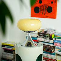 Die cremefarbene Space Age Lampe mit Chromfuß steht auf einem modernen Beistelltisch vor einem Bücherstapel. Im Hintergrund ein abstraktes, orangerotes Kunstwerk, das den Retro-Charakter der Szene unterstreicht.





