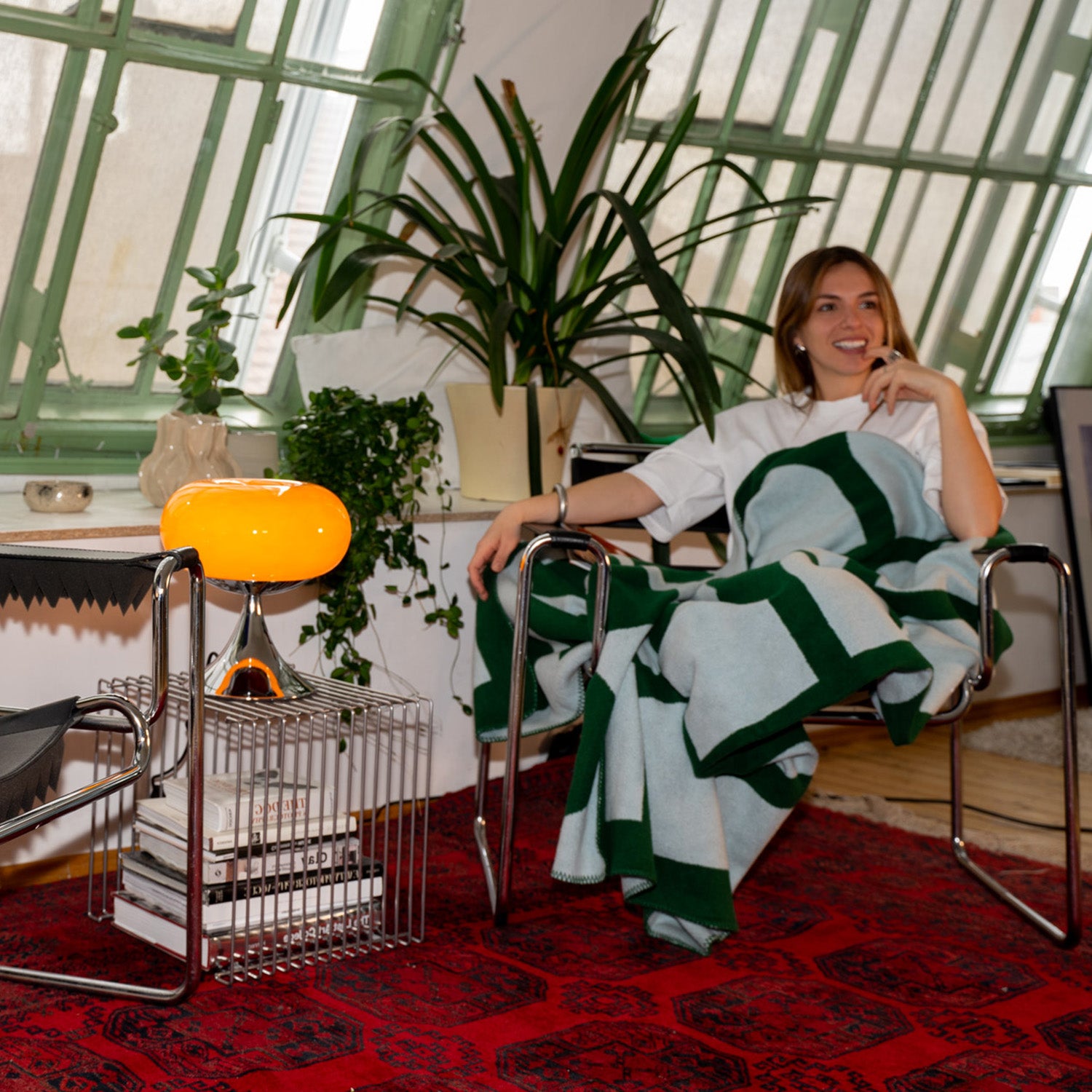 Gemütliche Ecke mit rotem Teppich und verchromtem Stuhl, in dem eine Frau mit weißem Shirt und grün-gemusterter Decke sitzt. Daneben ein Drahttisch mit Büchern und orangefarbener Midcentury-Lampe, umgeben von Pflanzen vor grüner Fensterfront.