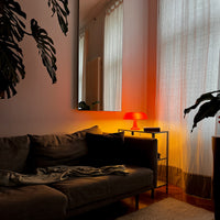 Die orange Vintage-Lampe auf einem Metall-Beistelltisch neben einem grauen Sofa mit dunklem Kissen. Der warme Lichtschein spiegelt sich im großen Wandspiegel wider, während transparente Vorhänge das Tageslicht sanft filtern.