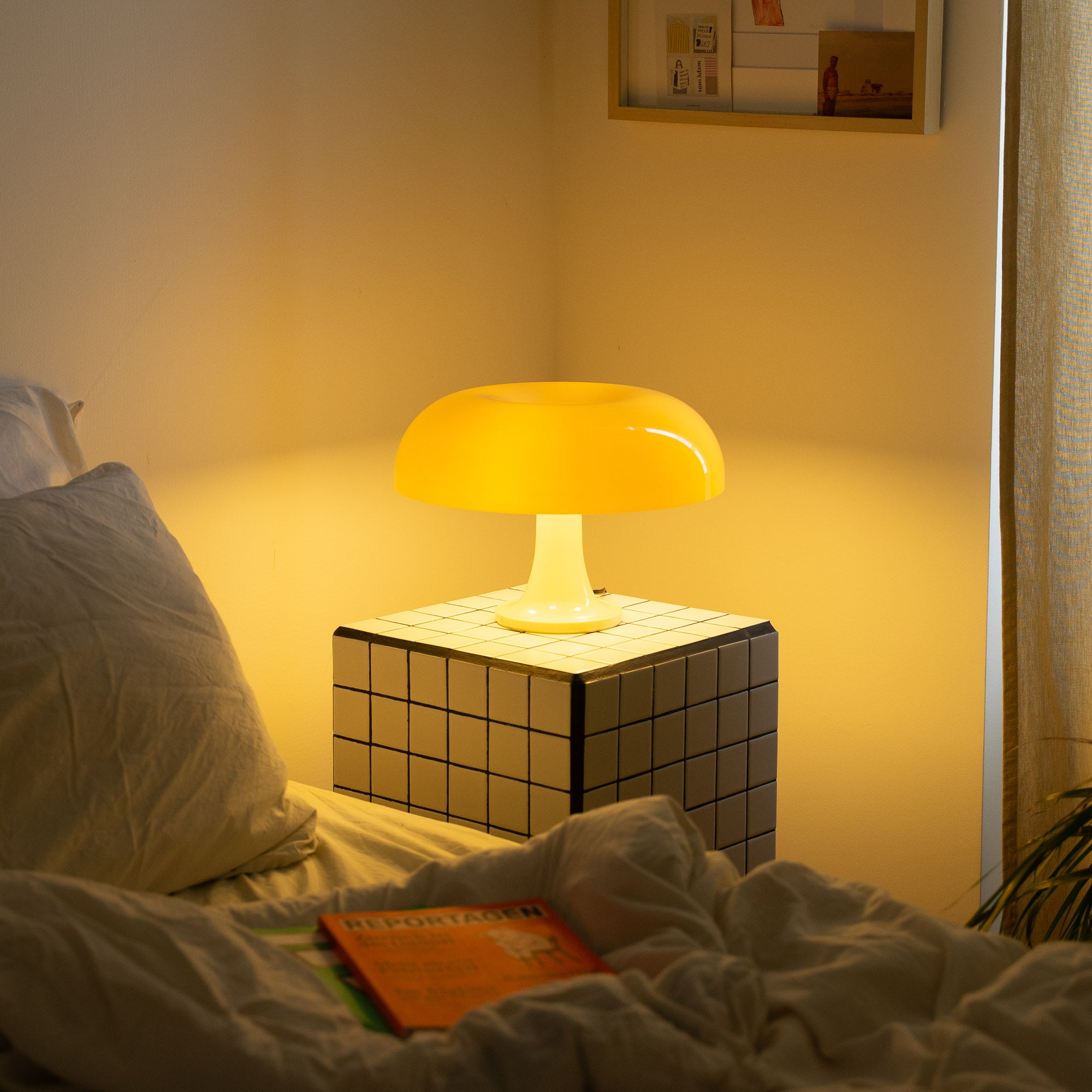 Die gelbe Tischlampe erhellt den Raum mit einem warmen Licht. Sie steht auf einem modernen Fliesentisch neben einem ungemachten Bett mit Magazinen. Ein cremefarbener Vorhang und dezente Wandkunst runden das gemütliche Ambiente ab.