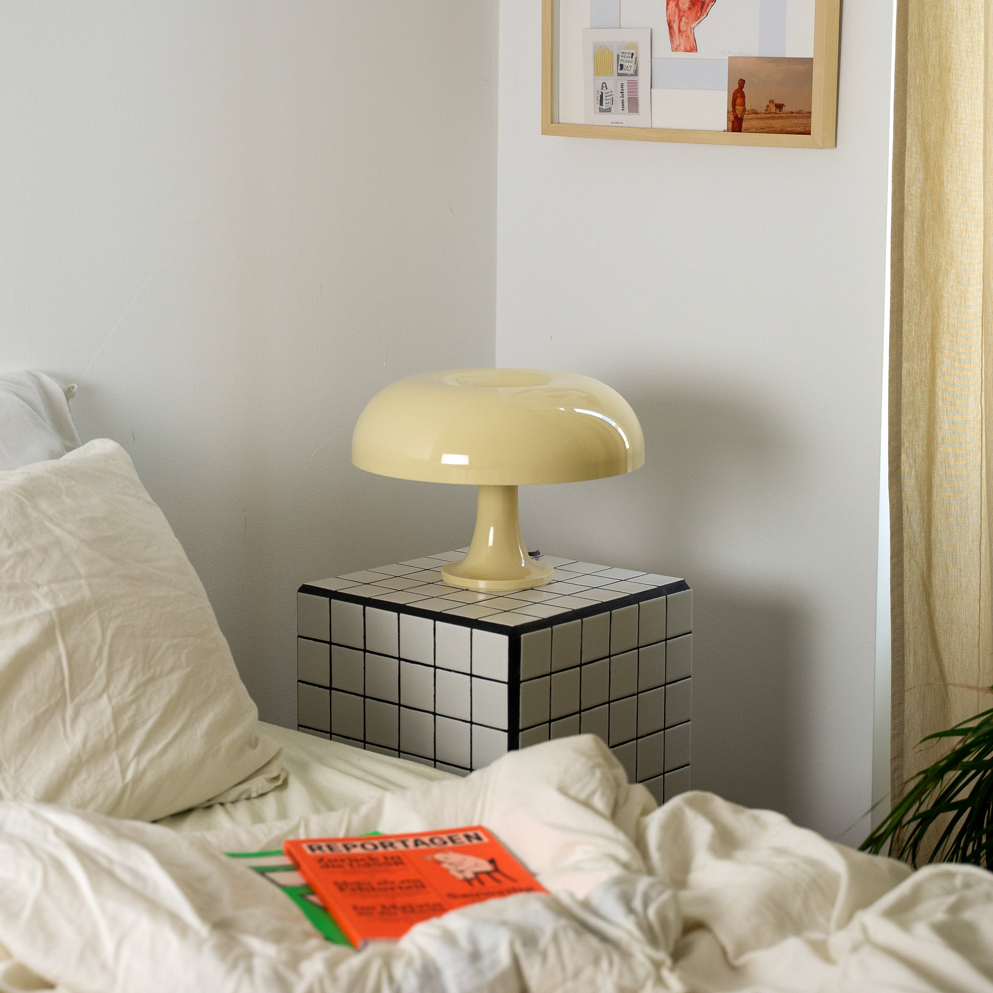 Die cremefarbene Pilzlampe steht auf einem schwarz-weiß karierten Beistelltisch neben einem Bett mit weißer Bettwäsche. Im Hintergrund eine minimalistische Wanddekoration und ein beiger Vorhang, der sanftes Licht hereinlässt.