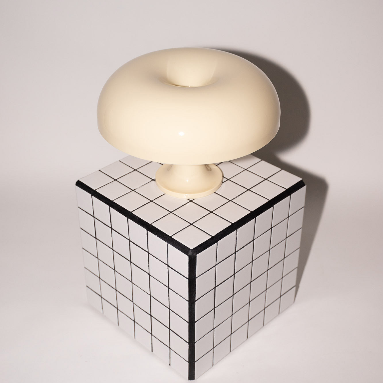 Die cremefarbene Acryl-Pilzlampe thront auf einem schwarz-weiß karierten Würfeltisch vor einer schlichten, hellen Wand. Die klare Linienführung und das glänzende Finish unterstreichen den Mid-Century Charme der Leuchte.