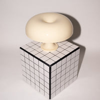Die cremefarbene Acryl-Pilzlampe thront auf einem schwarz-weiß karierten Würfeltisch vor einer schlichten, hellen Wand. Die klare Linienführung und das glänzende Finish unterstreichen den Mid-Century Charme der Leuchte.