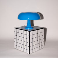 Die blaue Pilzlampe mit ihrem glänzenden Acrylschirm steht auf einem weiß-schwarz karierten Würfel-Beistelltisch vor einer neutralen Wand. Die klare Linienführung und das moderne Design unterstreichen den Mid-Century-Charakter.