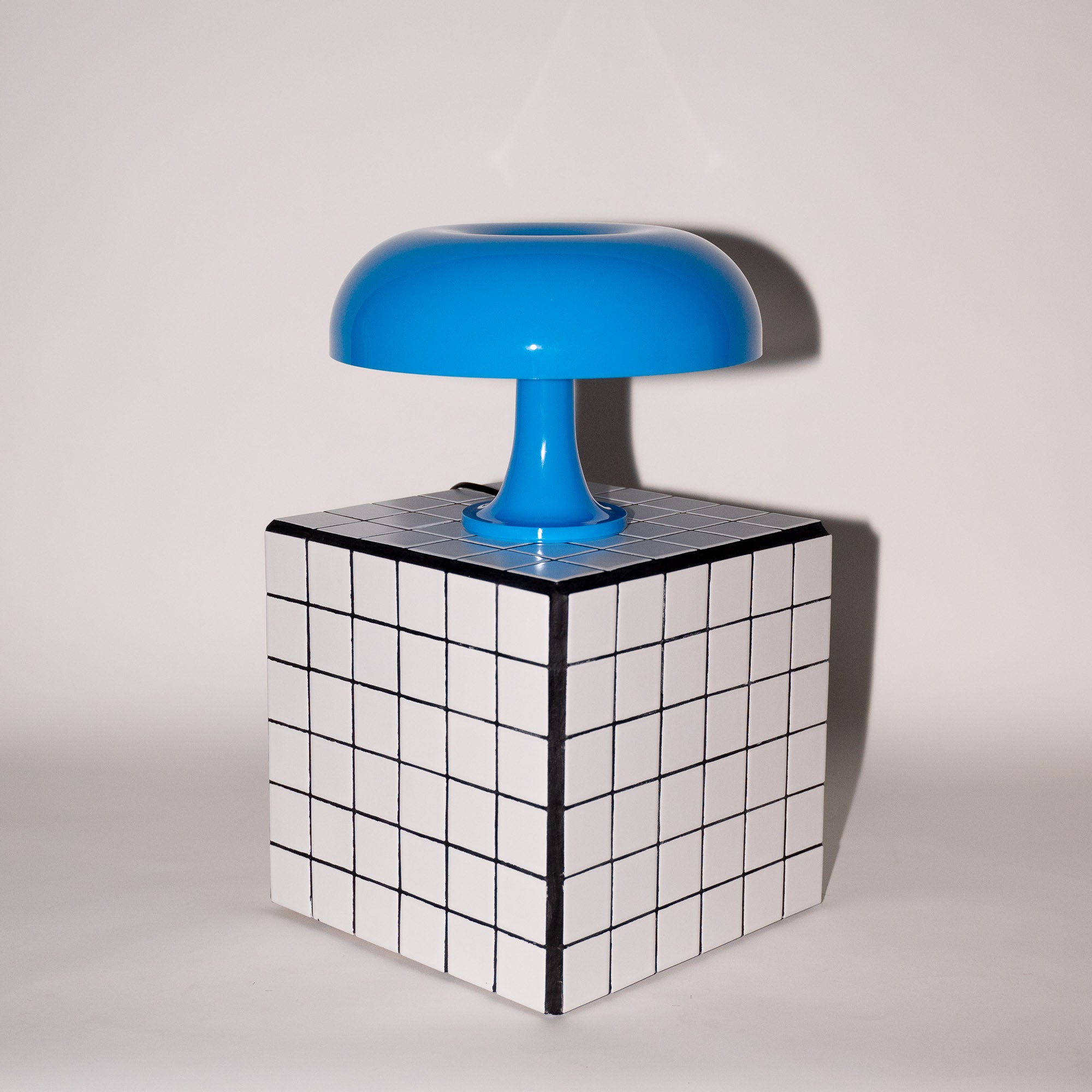 Die blaue Pilzlampe mit ihrem glänzenden Acrylschirm steht auf einem weiß-schwarz karierten Würfel-Beistelltisch vor einer neutralen Wand. Die klare Linienführung und das moderne Design unterstreichen den Mid-Century-Charakter.