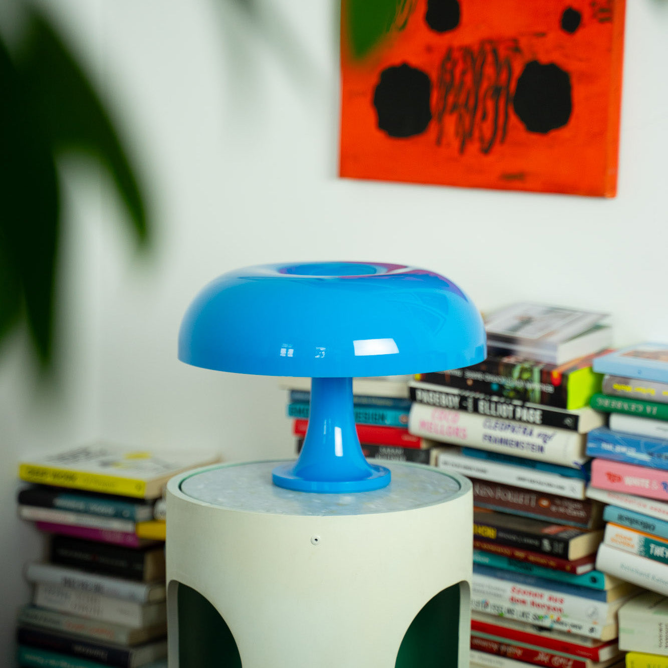 Die glänzende blaue Acryl-Tischlampe steht auf einem runden Beistelltisch, umgeben von bunten Büchern. Ein modernes Kunstwerk in kräftigem Orange ergänzt die Retro-Ästhetik des Raumes.