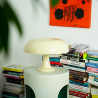 Die cremefarbene Tischlampe in Form eines Pilz steht auf einem runden Beistelltisch, umgeben von bunten Büchern. Ein modernes Kunstwerk in kräftigem Orange ergänzt die Retro-Ästhetik des Raumes.