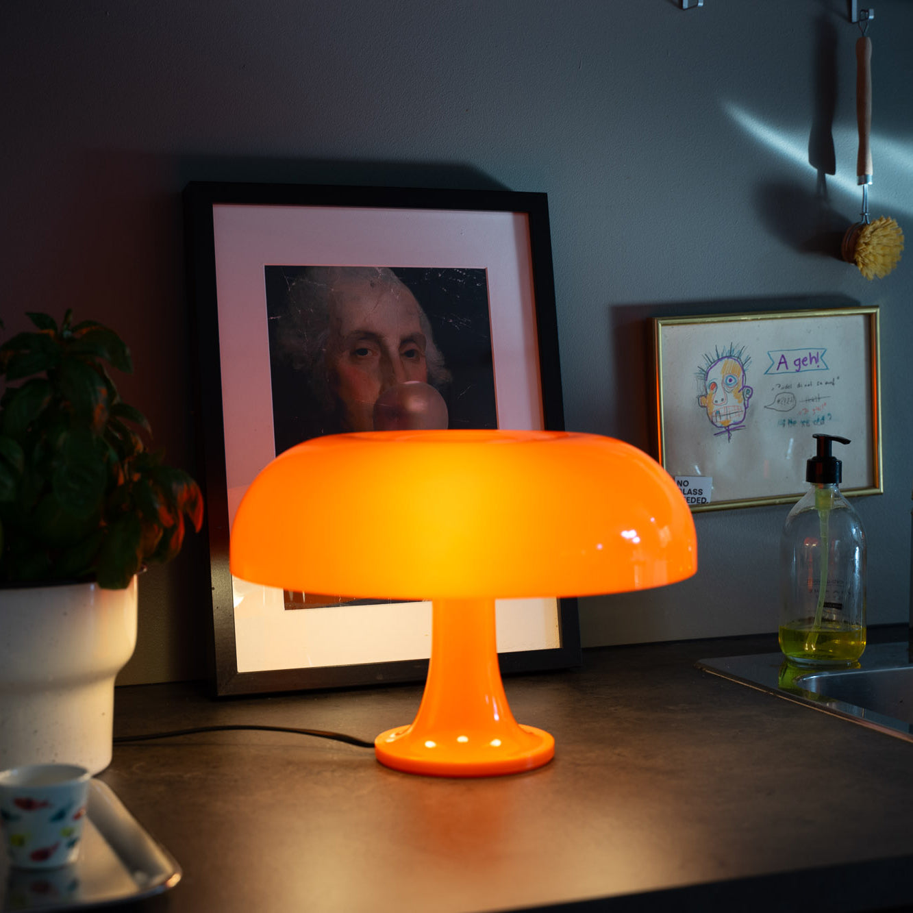 Die orange Pilzlampe steht auf einer dunklen Arbeitsplatte und taucht den Raum in warmes Licht. Dahinter ein gerahmtes Kunstwerk mit humorvollem Motiv, eine Zeichnung und Küchenutensilien, die eine kreative Atmosphäre schaffen.