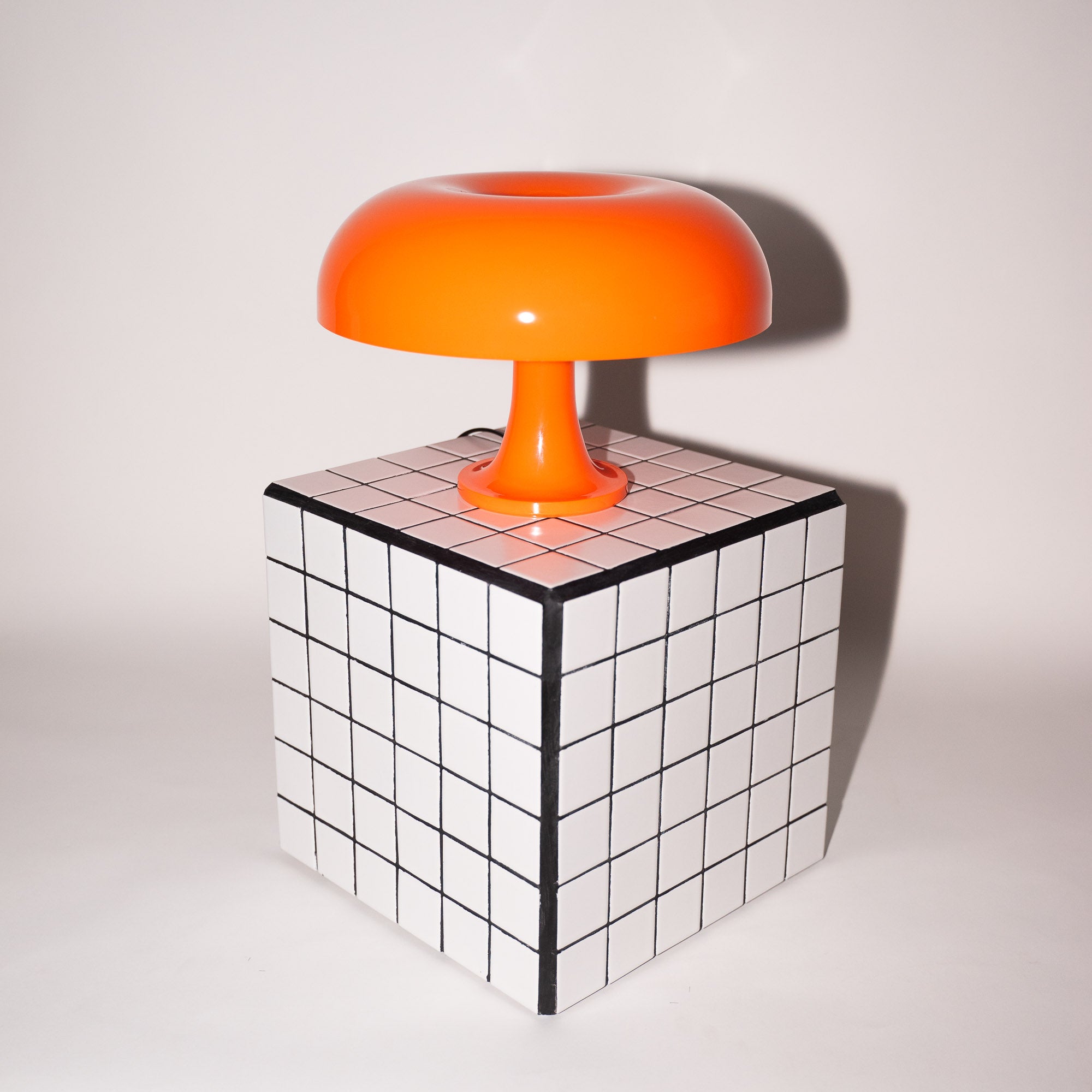 Die orange Pilzlampe steht auf einem schwarz-weiß karierten Würfeltisch vor hellem Hintergrund. Die klare Linienführung des Tisches verstärkt die futuristische Form der Lampe und betont ihren Mid-Century Charme.