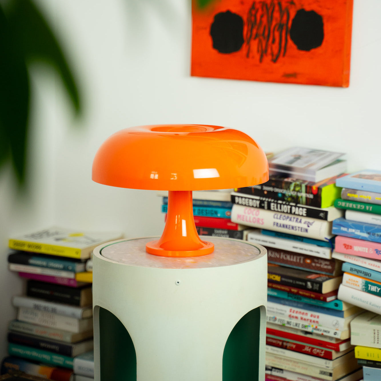 Die orange Acryl-Pilzlampe thront auf einem modernen runden Beistelltisch vor einer Wand mit abstrakter Kunst. Dahinter ein hoher Bücherstapel, der der Szene eine kreative und wohnliche Atmosphäre verleiht.
