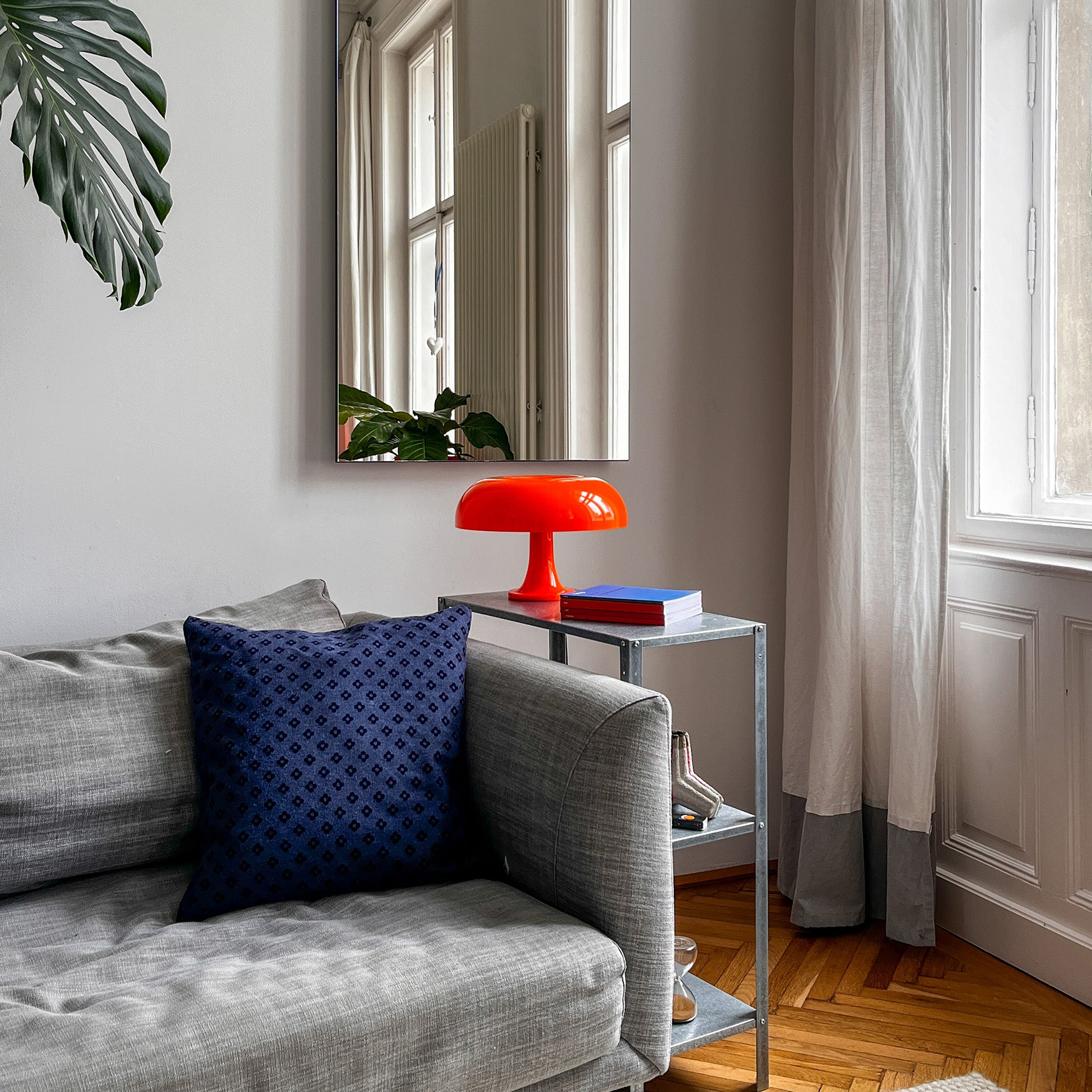 Die orange Space Age Lampe steht auf einem schlichten Metall-Beistelltisch neben einem grauen Sofa mit dunkelblauem Kissen. Große Fenster mit leichten Vorhängen bringen Tageslicht in den modern eingerichteten Raum.