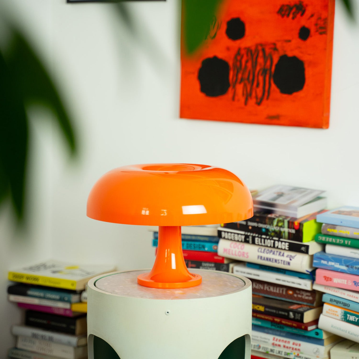 Die orange Acryl-Pilzlampe thront auf einem modernen runden Beistelltisch vor einer Wand mit abstrakter Kunst. Dahinter ein hoher Bücherstapel, der der Szene eine kreative und wohnliche Atmosphäre verleiht.