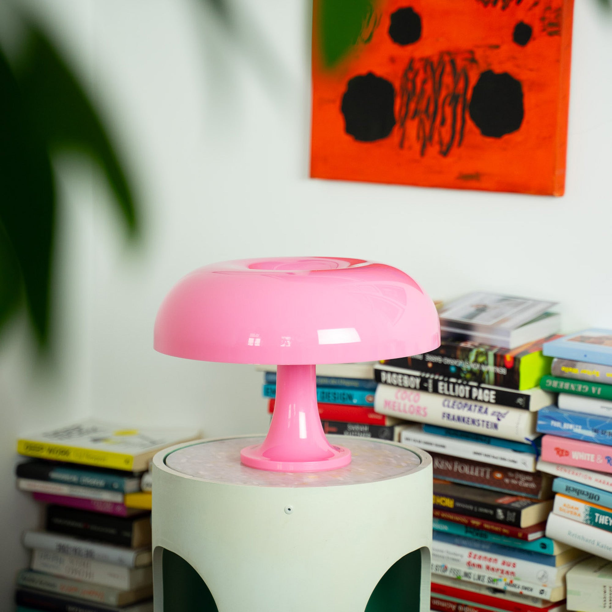  Die rosa Mid-Century Lampe steht auf einem runden Vintage-Sideboard vor einem Bücherstapel. Das kräftige Pink setzt einen verspielten Akzent in der modernen Wohnraumgestaltung.
