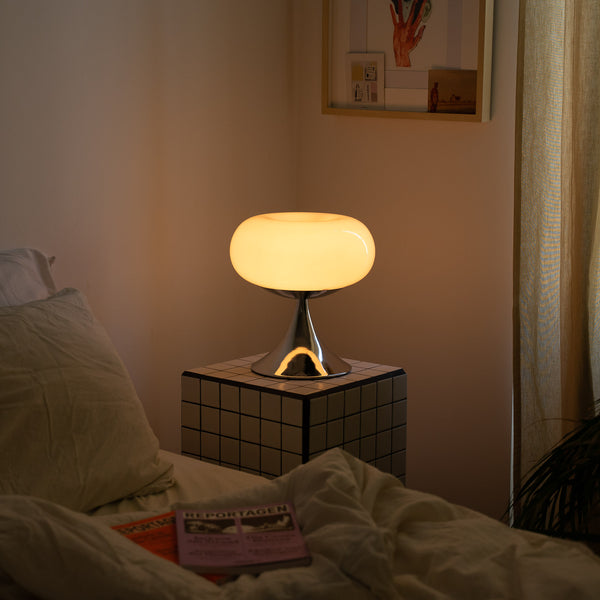 Gemütliche Nachtszene mit einer Space Age Tischlampe auf einem schwarz-weiß karierten Beistelltisch. Die Lampe mit opalem Glasschirm und Chromfuß strahlt warmes Licht aus. Daneben ein ungemachtes Bett mit Magazinen, helle Vorhänge und eine minimalistische Wandgestaltung.