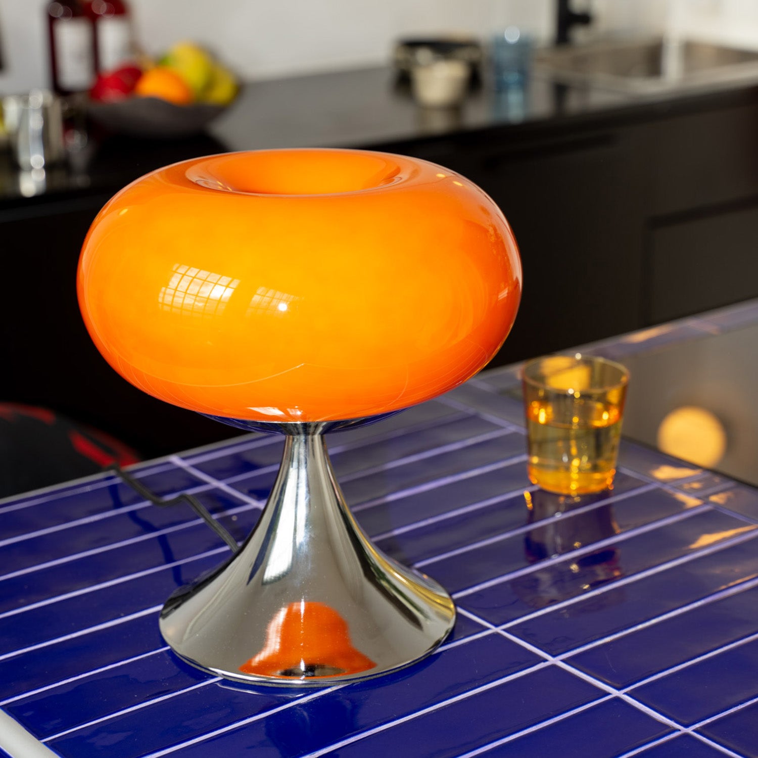 Eine auffällig geformte Tischlampe mit leuchtend orangefarbenem Glasschirm und glänzendem Chromfuß steht auf einer dunkelblau gekachelten Kücheninsel. Im Hintergrund sind eine schwarze Küchenzeile, ein Waschbecken sowie eine Obstschale und ein Trinkglas sichtbar. Das Design der Lampe erinnert an Mid-Century-Stil und setzt einen farbenfrohen Akzent im modernen Küchenambiente.