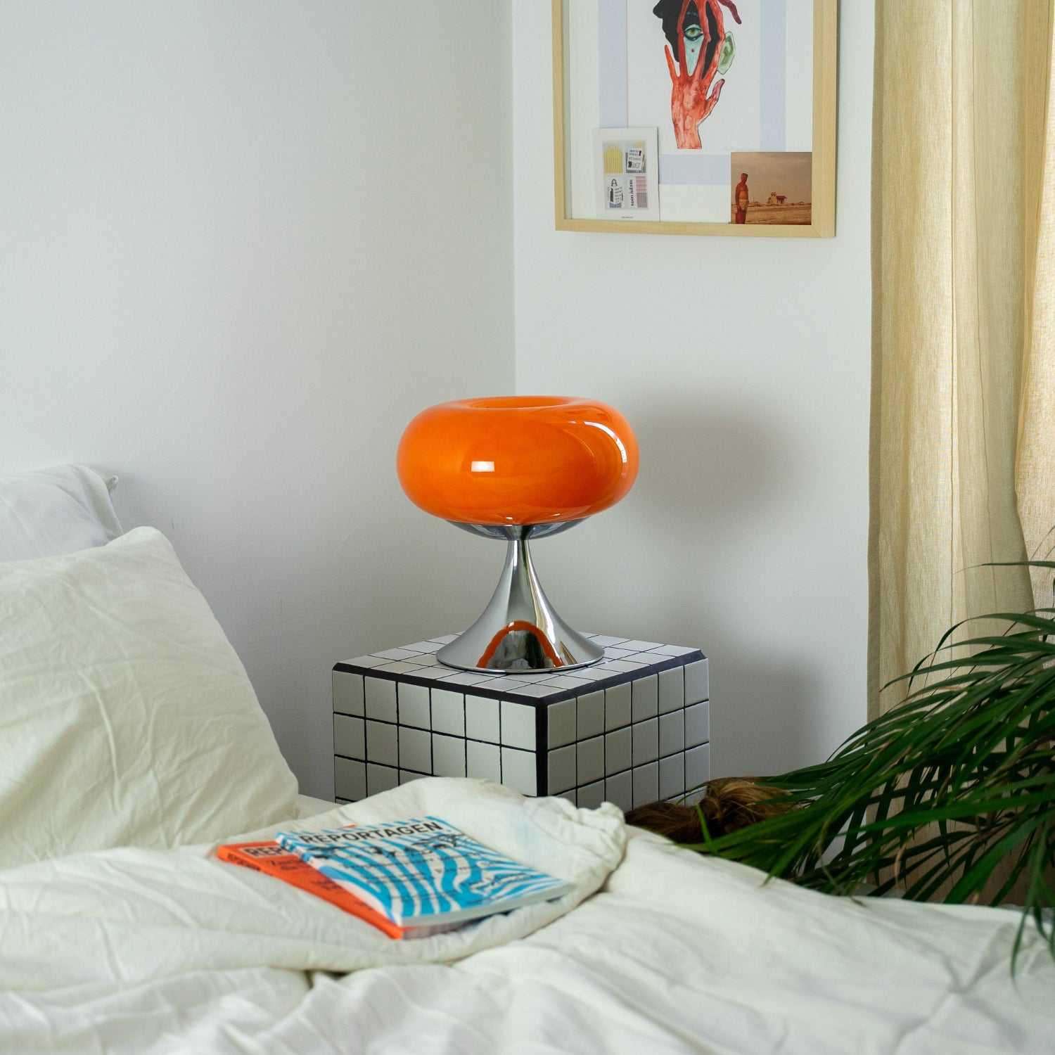 Die Mid-Century-Lampe steht auf einem karierten Beistelltisch neben einem Bett mit beiger Bettwäsche. Eine Zeitschrift liegt auf der Decke, sanftes Tageslicht fällt durch helle Vorhänge.