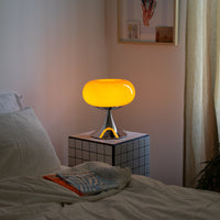 Die orangefarbene Space Age Tischlampe steht auf einem karierten Beistelltisch neben einem Bett mit weißer Bettwäsche. Die sanfte Beleuchtung schafft eine beruhigende Atmosphäre. Eine Zeitschrift liegt locker auf der Decke.