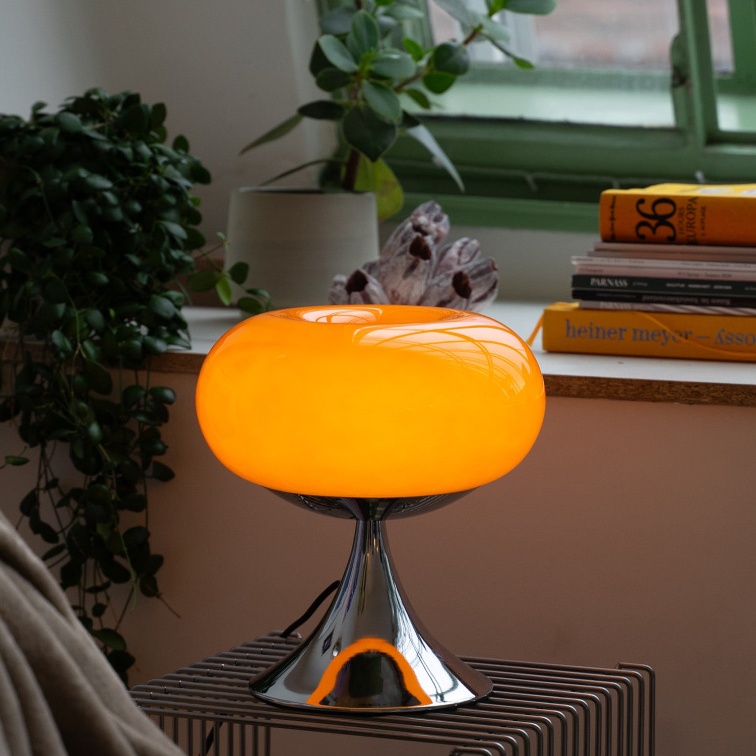 Orange Donut Lampe von GLOW UP STUDIO mit glänzendem Chromfuß steht auf einem gerillten Metall-Beistelltisch vor einem Fenster mit grünem Rahmen. Die Lampe strahlt ein warmes Licht aus, das den gemütlichen Wohnbereich mit Zimmerpflanzen, Kunstobjekten und einem Stapel Bücher auf der Fensterbank sanft beleuchtet. Die Szene wirkt stilvoll und einladend.