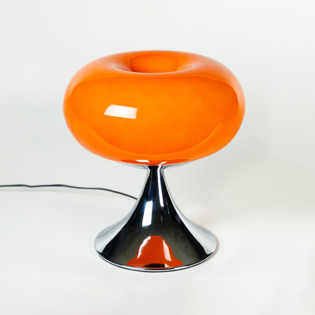 Die glänzende Glasleuchte mit rundem, organischem Design und trompetenförmigem Chromfuß erinnert an das ikonische Space Age Design der 60er und 70er Jahre. Ideal als dekorative Nachttisch- oder Wohnzimmerlampe.