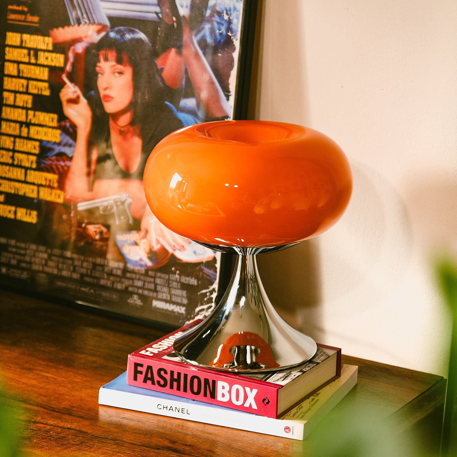 Orange Donut Lampe von GLOW UP STUDIO – Mid Century Lampe mit Chromfuß auf Bücherstapel im Wohnzimmer mit Pulp Fiction Filmplakat im Hintergrund