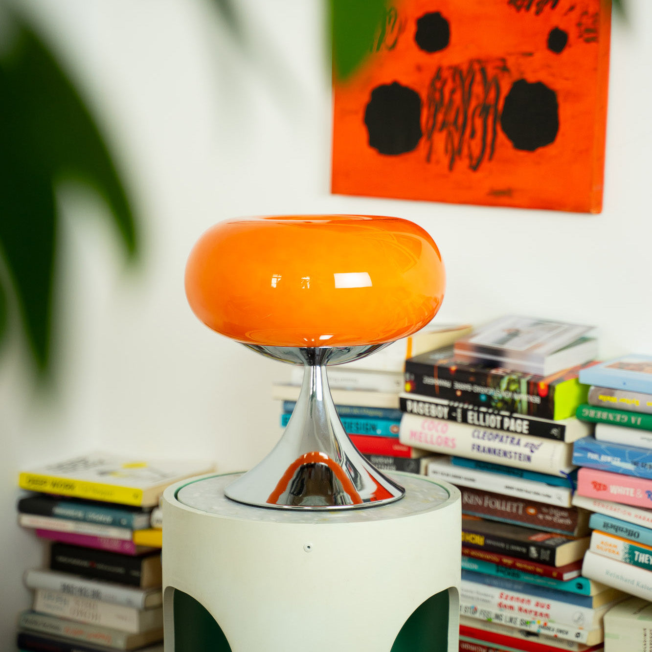 Die orange Glaslampe mit Chromfuß steht auf einem modernen Beistelltisch vor einem Bücherstapel. Im Hintergrund ein abstraktes Kunstwerk mit leuchtenden Farben, das die 70er-Jahre-Ästhetik der Lampe unterstreicht.